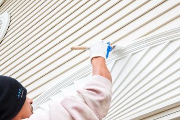 A worker applying white paint with a brush to the outside of a home.