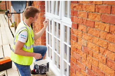 A worker painting white window trim in the back of an house.