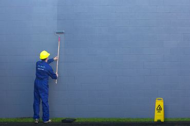 A man standing in front of a concrete wall using a roller brush to apply paint.