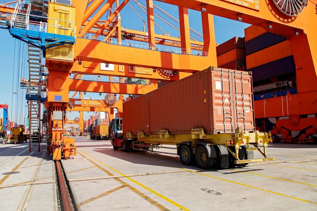 A truck carrying a large shipping container at a busy port with orange cranes.