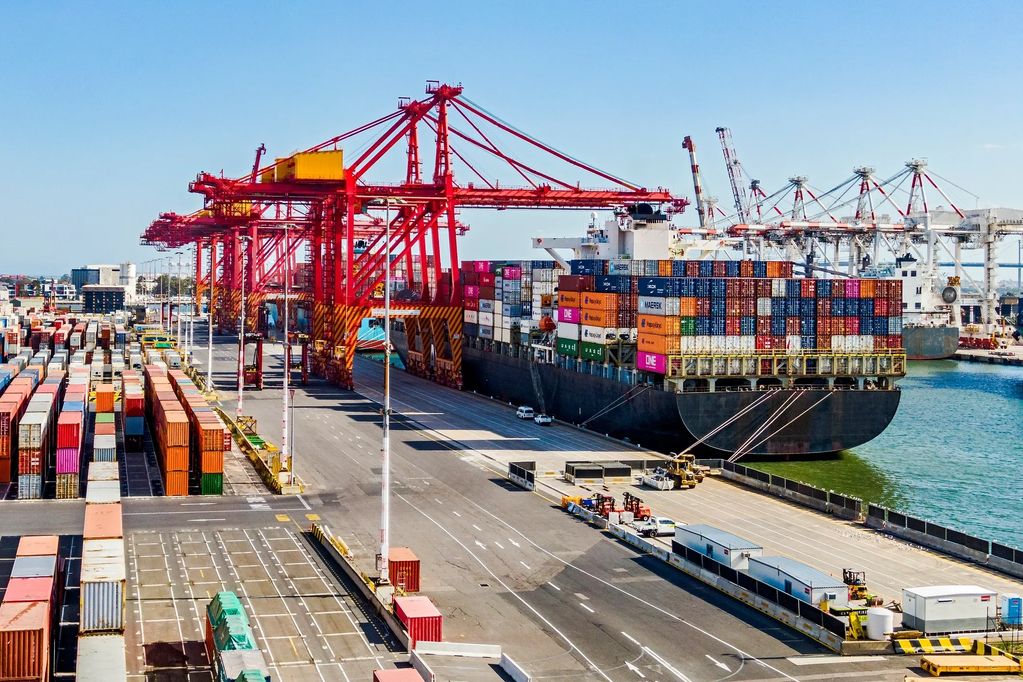 Cargo ship docked at port with red cranes and stacked containers.