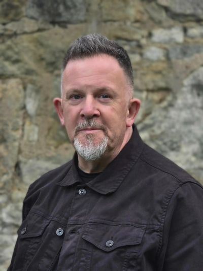 Man with a beard in a black jacket standing outdoors.
