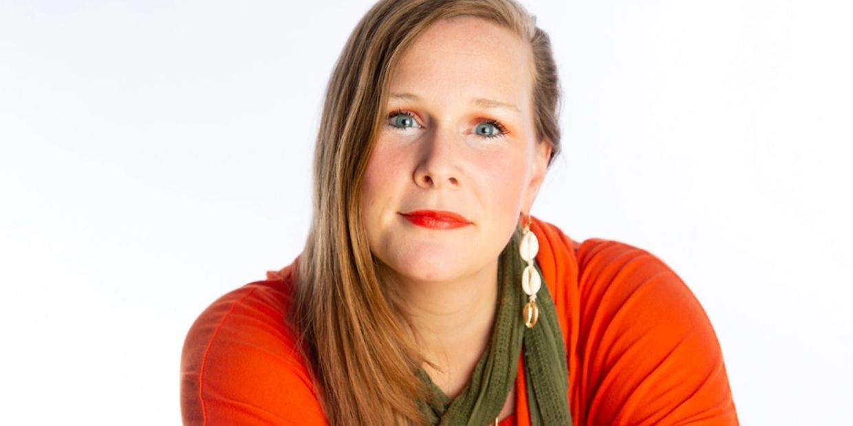 Woman with blue eyes, red lipstick, orange top, and shell earrings looks confidently at the camera.