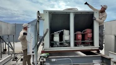Two workers repair a rooftop HVAC unit under a cloudy sky.