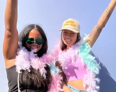 Two women smiling, wearing colorful feather boas and posing joyfully.