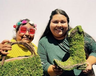 Two women smiling and holding moss-covered fashion items.
