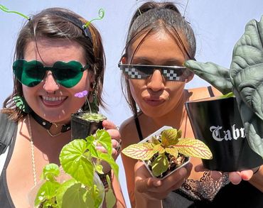 Two women holding potted plants, wearing fun sunglasses and smiling.
