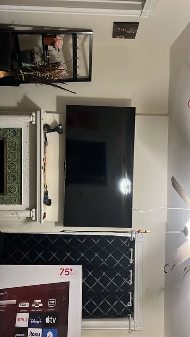 A mounted 75-inch TV above a fireplace with tools and a window with curtains.