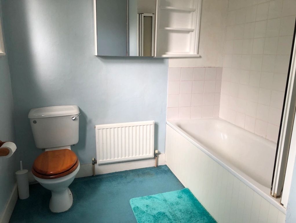 A simple bathroom with blue carpet, wooden toilet seat, and white bathtub.