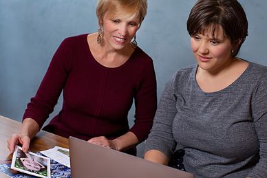 Two women smiling at a computer, one holding a brochure