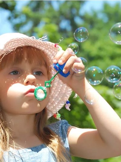 Child blowing bubbles with a pink straw hat and trees in the background at a festival