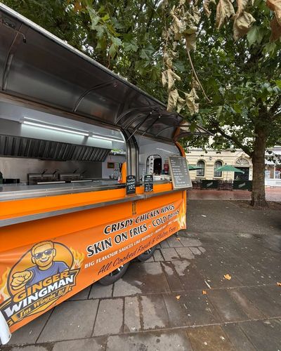 The bright orange Ginger Wingman trailer pitched under trees, serving hatch open