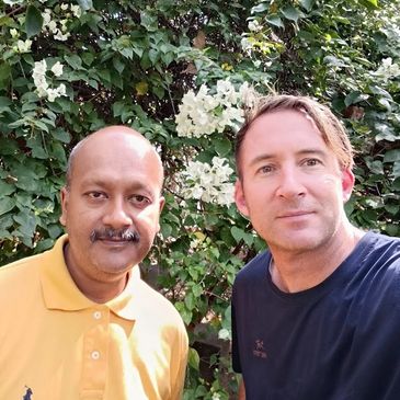 Two men posing for a selfie in front of lush greenery and white flowers.