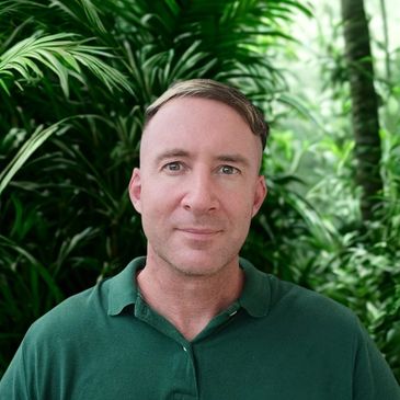 A man in a green shirt stands before lush tropical plants.