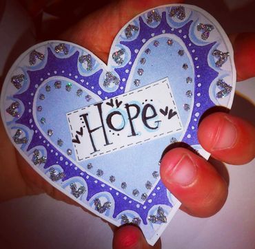 A hand holding a decorated heart-shaped paper with the word 'Hope'.