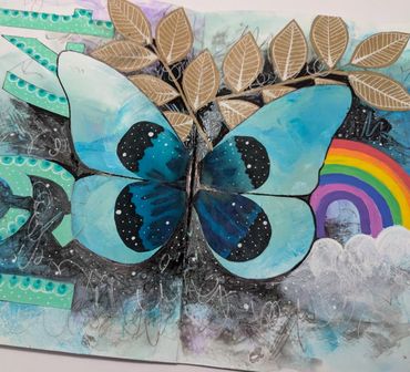 Artistic butterfly with blue wings, leaves, and a rainbow on a mixed media background.