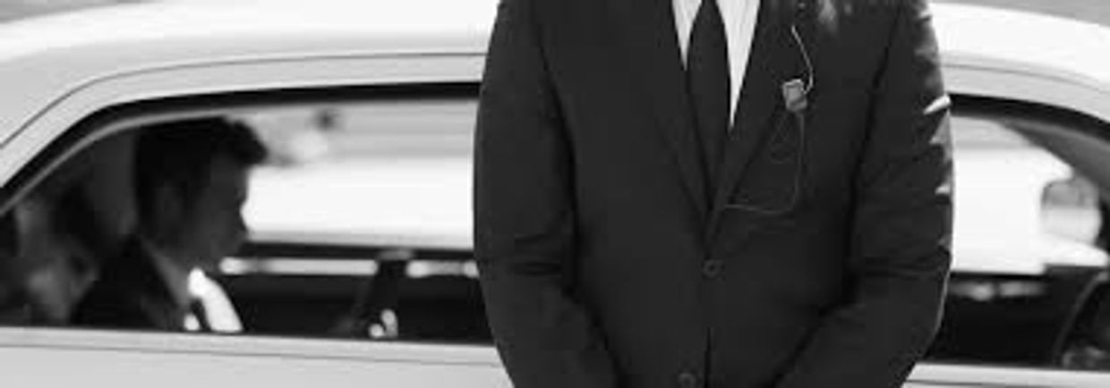 A suited man standing in front of a car with a passenger inside.