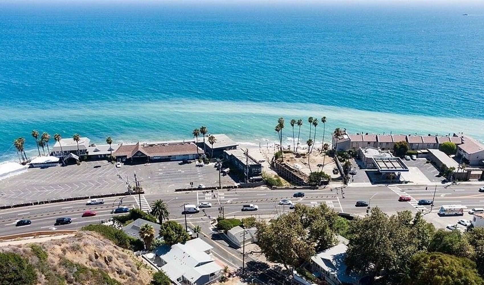 A coastal road with cars beside a turquoise ocean and palm trees.