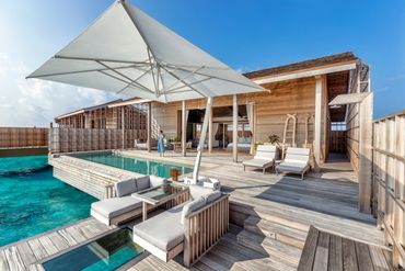 Luxury wooden overwater villa with pool, lounge chairs, and large umbrella under clear blue sky.