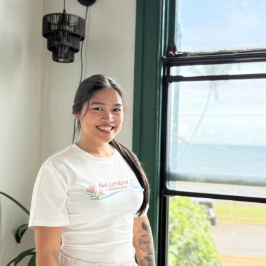Smiling woman with long dark hair standing by a window with a view of the ocean.