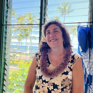 Smiling woman in a floral dress with a lei and flower in her hair near a window with palm trees outside.