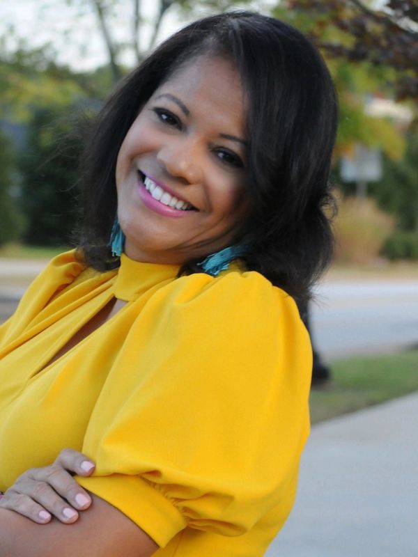 Woman smiling outdoors wearing a bright yellow top and turquoise earrings.