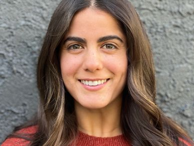 Smiling woman with long brown hair in a red sweater against a gray textured wall.