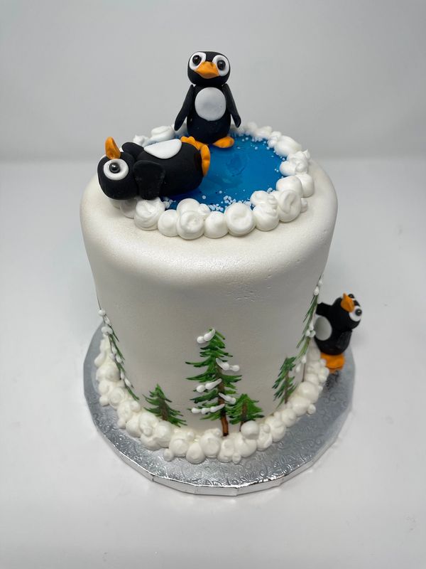White cake with ice pond and penguins on top, hand painted pine trees on side.
