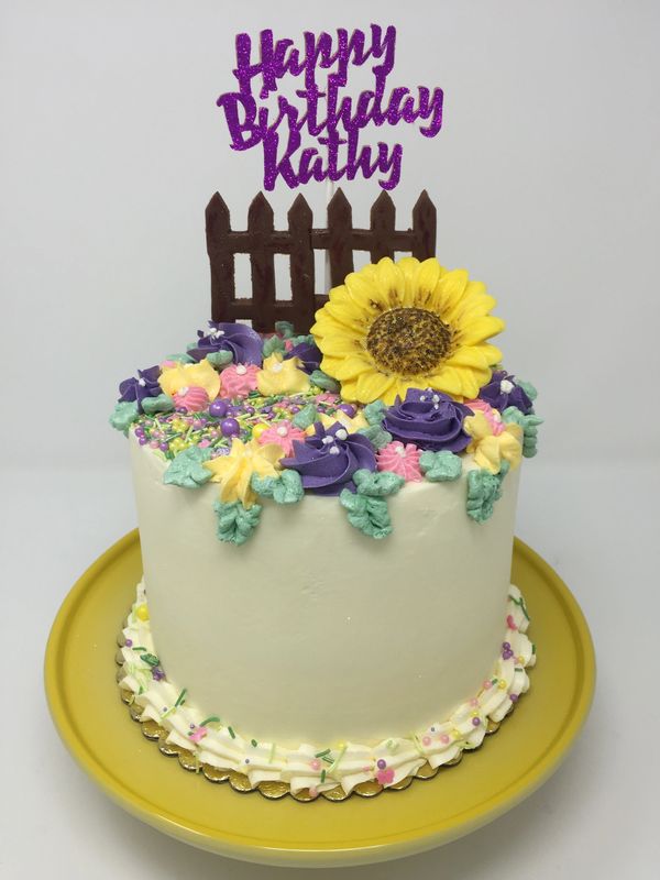 white cake with sunflower and brown fence on top surrounded by multicolored 