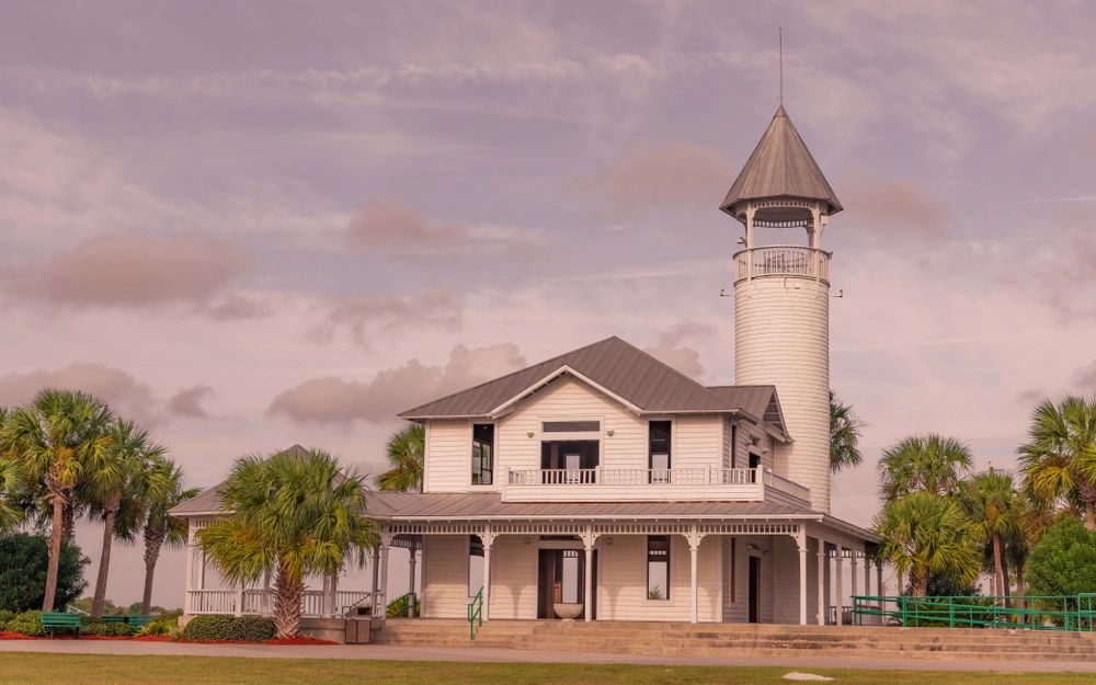 Historic white lighthouse building surrounded by palm trees under a cloudy sky.