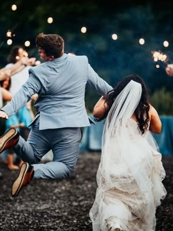 Groom joyfully jumps as bride walks through sparklers held by guests.