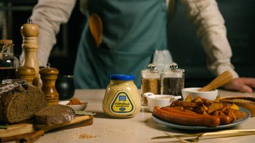 Jar of mustard on kitchen counter with bread, sausages, and spices.