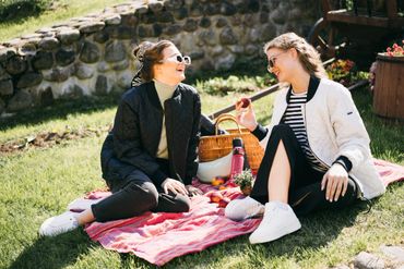 Two women enjoying a sunny picnic on a red blanket outdoors.