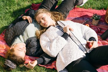 Two women relaxing on a picnic blanket in a sunny park, one holding an apple.