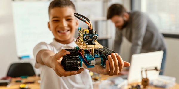 Boy proudly shows a homemade robot with tank tracks in a classroom.