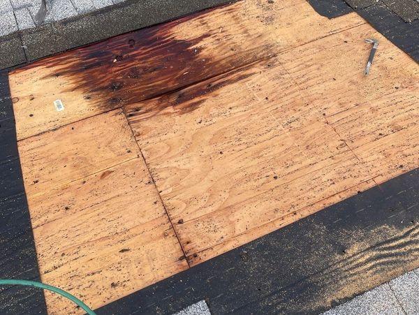Roof repair in progress showing replaced plywood decking due to water damage beneath removed shingles, with roofing tools on site.