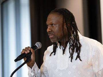 Man with dreadlocks speaking into a microphone indoors.