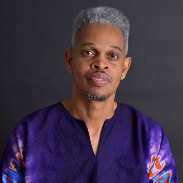 Man with gray hair wearing a purple traditional outfit against a dark background.