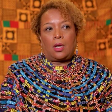 Woman speaking, dressed in colorful traditional African attire with patterned background.
