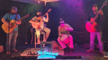 A four-member band performing on stage with guitars and a drum at The Doghouse.