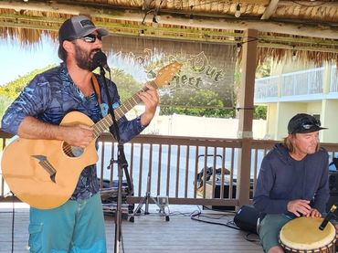 Two musicians perform on a wooden stage under a thatched roof.