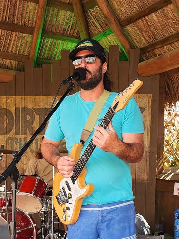 Man playing electric guitar and singing under a thatched roof stage.