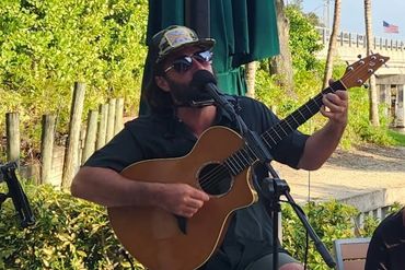 Man playing guitar and harmonica outdoors with sunglasses and cap.