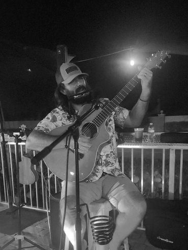 Man playing guitar and harmonica barefoot on a stool under purple stage lights.