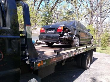 Damaged black Honda Civic on a tow truck bed in a sunny area.
