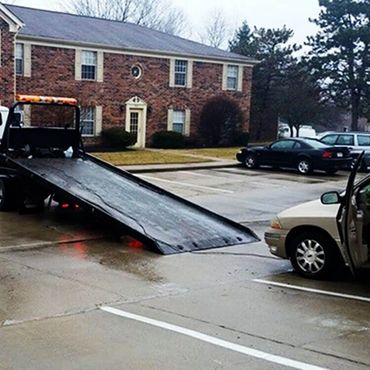 A tow truck with its ramp extended in front of a beige car in a wet parking lot.