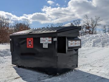 Black dumpster in a snowy outdoor area with cloudy sky.