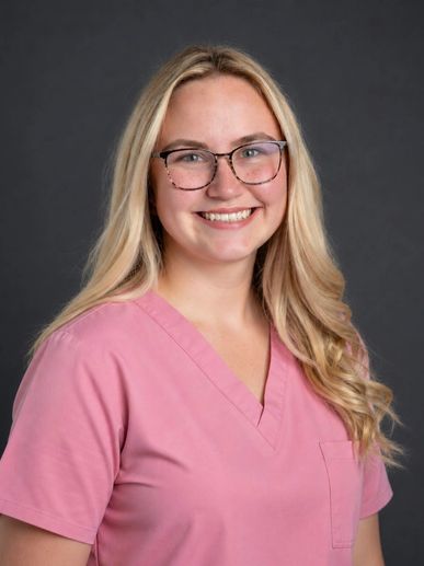 Smiling woman in pink scrubs with glasses against a dark background.