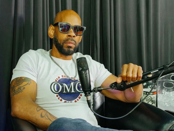 Man with sunglasses and tattoos speaking into a microphone in a studio.