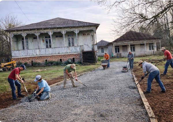 Group of people laying gravel on a rural driveway near old houses.